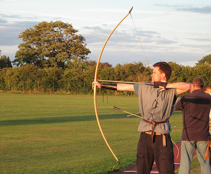 A historical reenactor in period kit drawing a longbow, showing the full extension and the height of the bow.